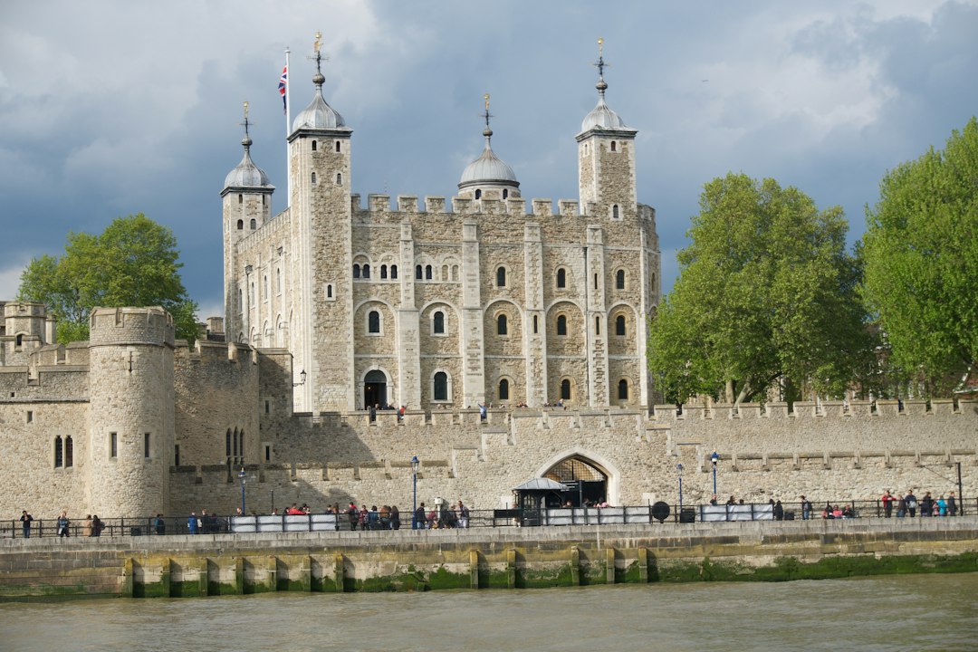 The Tower of London as viewed from the River Thames, its aged stone walls contrasting against a dramatic grey sky. The composition emphasizes the castle's imposing architecture, enhanced by the muted color palette and natural lighting. The presence of visitors subtly adds a sense of scale and historical context.